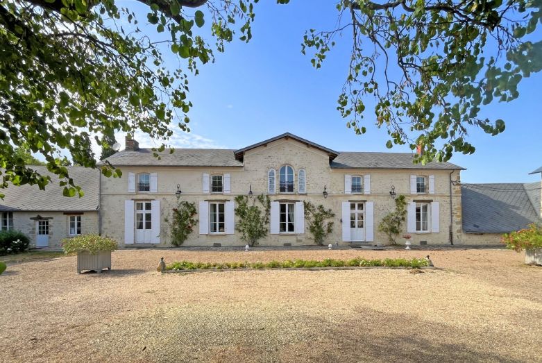 La façade de La Maison, pierre blonde et volets blancs, sous les arbres